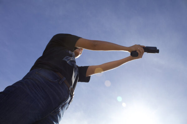 Student of NRA Basic Pistol Shooting course firing a handgun, as viewed from the ground, back-dropped by a sunny blue sky.