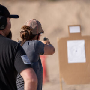 A wife shoots paper targets while her husband looks on, in Ministry of Defense Enhanced Concealed Weapons License class.